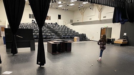 A photo of Highland Hall before renovations shows a person standing on the former theater stage, looking out to a closed room with auditorium seating and dropped ceiling.