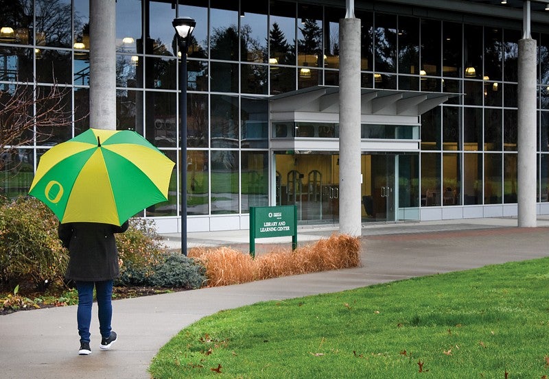 Student with green and yellow striped umbrella walking into Library and Learning Center at University of Oregon Portland.