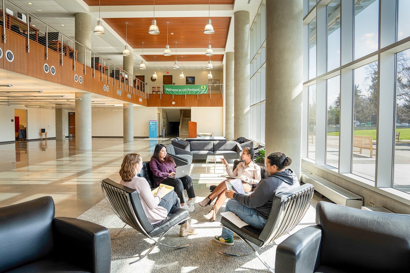 Four UO Portland employees meet in the open space of the Library and Learning Center. They are facing each other with a large wall of windows overlooking the outdoor green. 