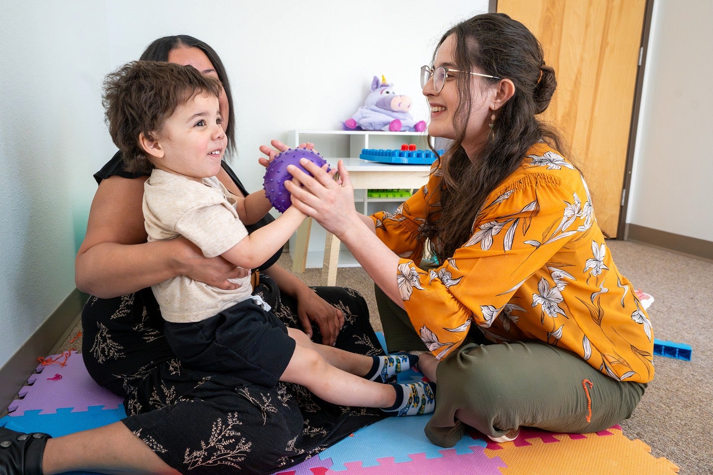 A student plays with a toddler who is sitting on a caregiver's lap.