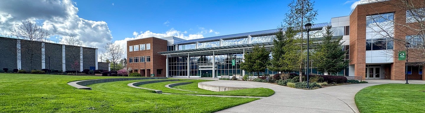 Outside of the Library and Learning Center on the UO Portland campus. A pathway splits to a Y, with the left path going to a small amphitheater in the grass and the right path going out of the photo. 