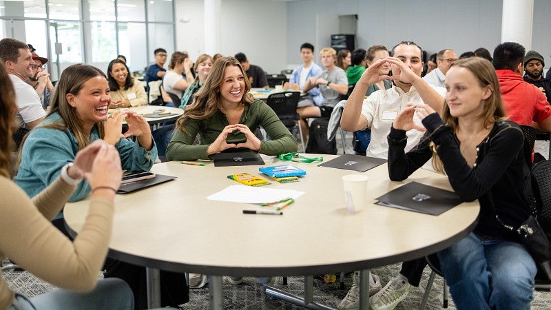 Students sit at a round table and hold up their hands in an O shape