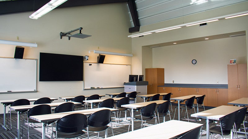 Classroom features rows of tables and chairs, two white boards, a display screen and counter space. 
