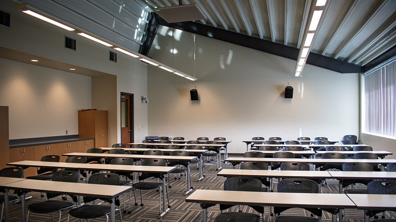Large classroom with rows of tables and chairs, vaulted ceiling, a wall of windows, and built in cabinets. 