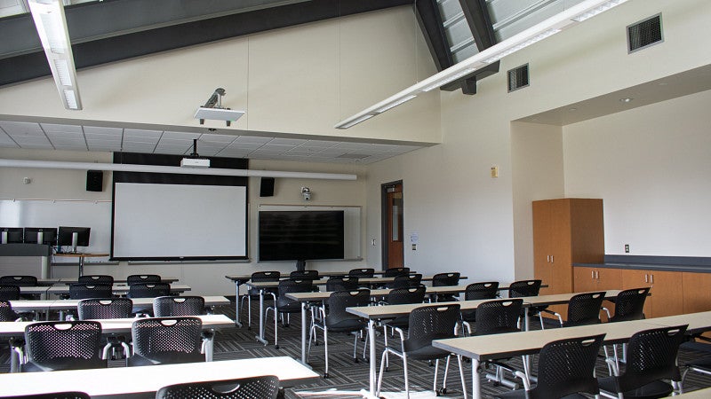 View of front of classroom with a projector screen, monitor screen and lectern. 