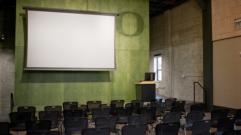 Rows of chairs in front of a stage with a projector screen, podium and O on the wall. 