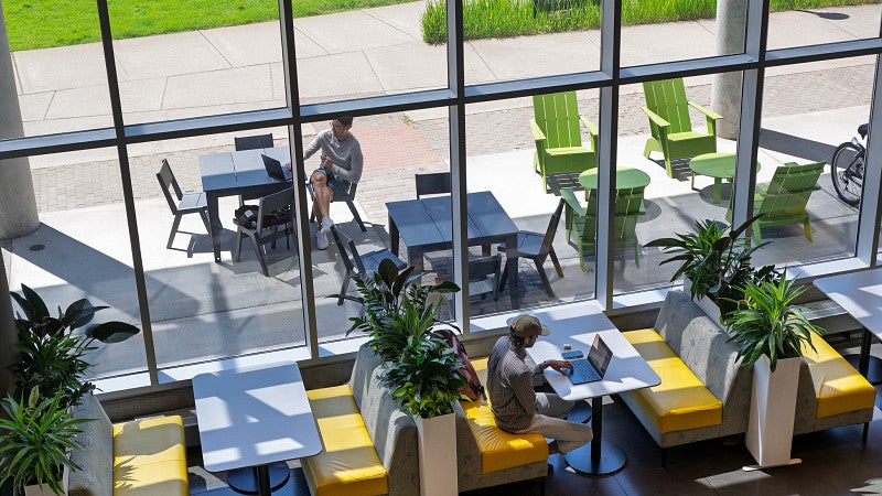 A person sits at a booth working on a laptop next to a wall of windows. Another person is seen sitting at a table outside. 