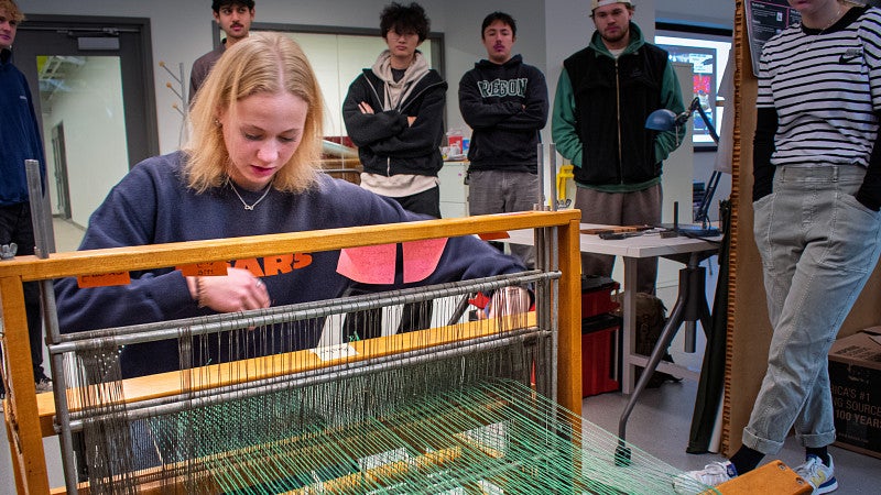 A student practices on a loom while others watch. 