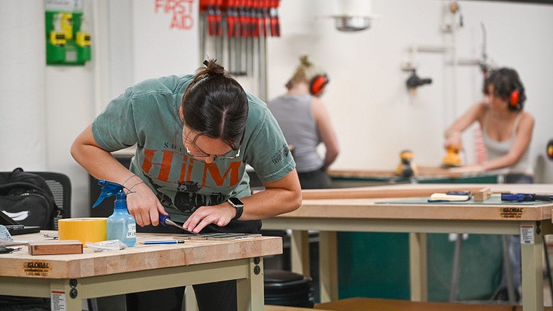 A student leans over a table in the wood shop to use pliers on a project. Other students sand wood in the background. 