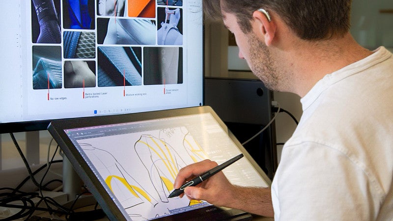 A product design student sketches on a tablet with a computer with images set up behind. 