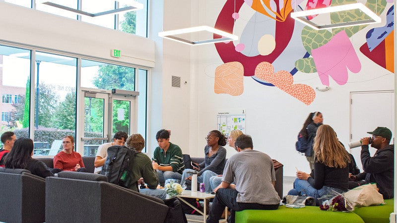 Students gather together in the Campus Center. A large art mural is on the wall in background. 