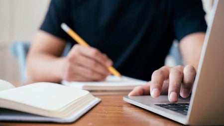 A person sits at a computer and writes on a notepad with a pencil. A book is open nearby. 