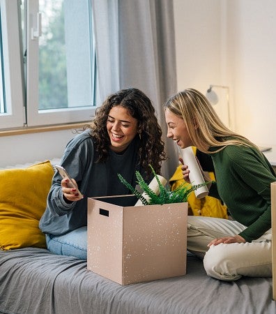 two roommates laugh at something on their phone. Moving boxes are placed around them.