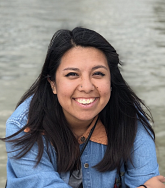 picture of Elizabeth Gillingham sitting in front of a body of water, smiling, with her arms crossed in front of her